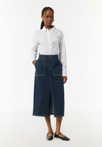 Jeans-Midi-Rock mit einem taillierten Bund, Schlitz vorne, zwei aufgesetzten Taschen, Kontrastnähten und in Dunkelblau. Dazu wird ein weißes Hemd getragen.