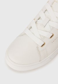 Baskets blanches en cuir avec surface texturée, présentant des accents dorés et des lacets blancs. Bout arrondi et semelle en caoutchouc plate. Nom de la marque visible.