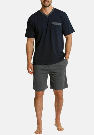 Man wearing a navy blue short-sleeve V-neck shirt with a gray chest pocket and gray knee-length shorts, barefoot.