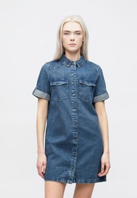 Denim skjortekjole med korte oppbrettede ermer, to frontlommer og knappestolpe foran. Har krage og frynset kant.