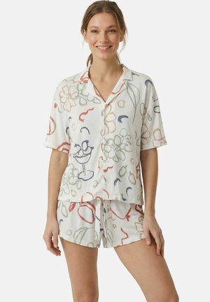 Jonge vrouw die een wit overhemd met korte mouwen en knopen draagt, samen met bijpassende shorts met een abstracte bloemen- en fruitprint, glimlachend naar de camera.