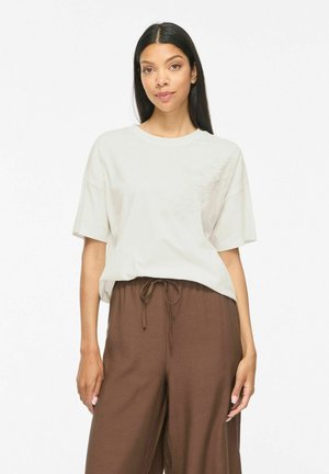 Femme aux longs cheveux foncés portant un haut blanc à manches courtes décontracté et un pantalon marron ample à cordon, debout devant un fond blanc.