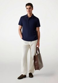 Polo bleu marine à manches courtes avec côtes verticales, pantalon léger crème, sac fourre-tout en cuir marron, et chaussures sans lacets marron foncé.