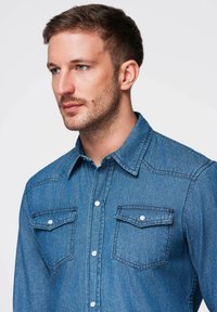 Chemise en denim bleu moyen avec un col avec boutons, manches longues, deux poches poitrine et des détails de quincaillerie argentée. Texture subtilement visible.
