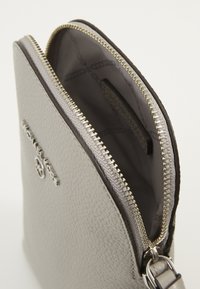 Grå strukturerad läderplånbok med silverblixtlås och metalllogotyp på framsidan, visar ett öppet inre fodrat med mönstrad tyg.