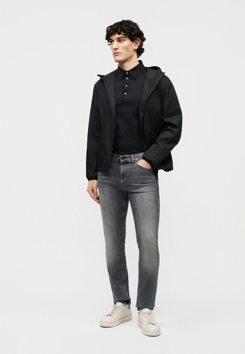 Giovane uomo in piedi, indossa una giacca con cappuccio nera, una polo nera con bottoni, jeans grigi sbiaditi e sneakers avorio.