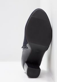 Chaussure à talon noire avec semelle texturée et fermeture éclair latérale, vue de dessous avec le talon appuyé contre une surface blanche.