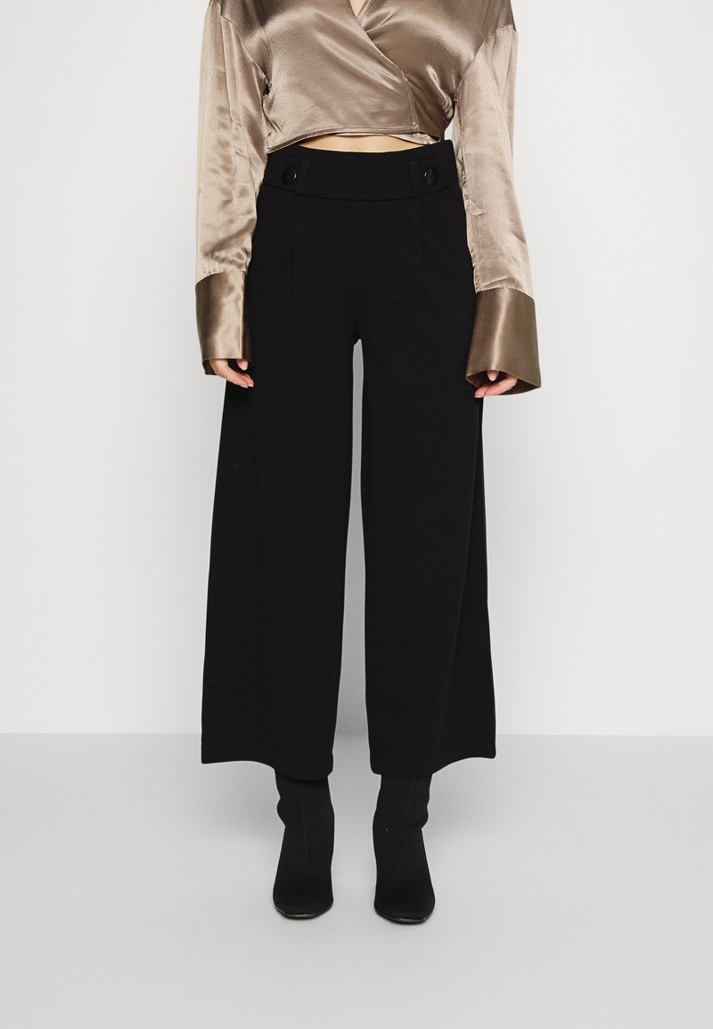 Femme portant un pantalon noir taille haute à jambes larges, associé à des bottines noires et un chemisier court brillant taupe à manches longues.