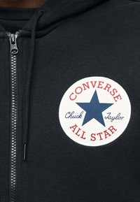 Μαύρο φούτερ με φερμουάρ και σ τρογγυλή επιγραφή που αναγράφει «CONVERSE ALL STAR», κόκκινα στοιχεία και μπλε αστέρι στο κέντρο. Μαλακή υφή υφάσματος.