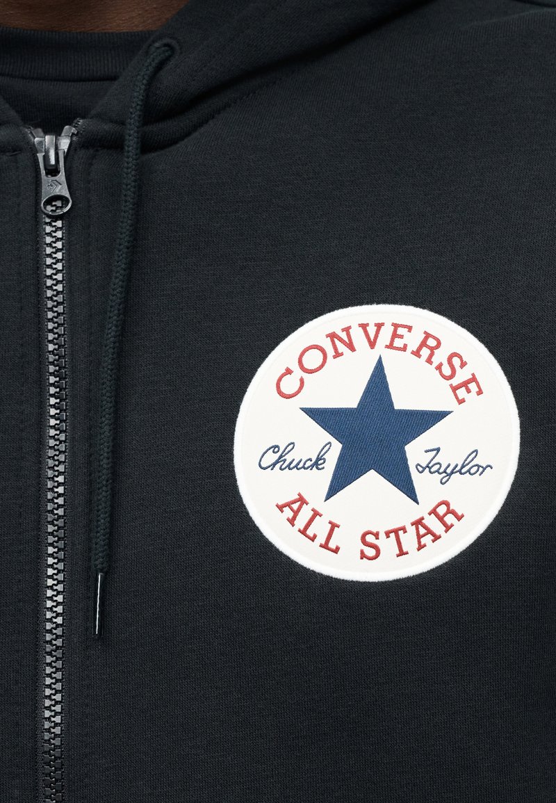 Sort hættejakke med lynlås, der har et rundt mærke med teksten "CONVERSE ALL STAR", røde detaljer og en blå stjerne i midten. Blød stoft struktur.