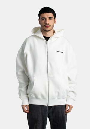 Joven con sudadera blanca con cremallera y logo "PEGADOR" en el pecho, pantalones negros, de pie frente a un fondo claro y liso.