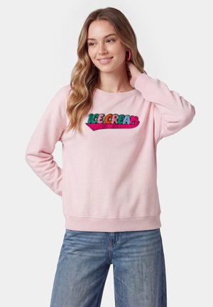Ung kvinde med langt hår iført en pink sweater med farverig "ICE CREAM" tekst og blå jeans, smilende med hånden på nakken.