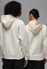 Beige-hupparit, joissa on kiristysnyörit, rento istuvuus ja resorireunukset, kaksi henkilöä pukeutuneena selkät vastakkain. Kangas on yksivärinen eikä siinä ole kuvioita.