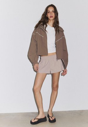 Ung kvinde iført brun cropped jakke, hvid crop top, beige shorts med sideliste og sorte platform klipklappere mod hvid væg.