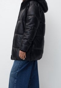 Doudoune noire avec capuche, présentant une surface lisse et brillante, des poignets élastiques et une silhouette carrée. Portée avec un jean bleu à jambes larges.