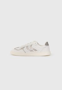Vit lågtoppsneaker med beige sula, perforerade sidopaneler, dekorativ sömnad och grå accent på hälfliken placerad på en enkel bakgrund.
