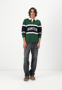 Grønn langermet polo med hvite og marineblå striper, med "HUNTER"-tekst over brystet; kombinert med grå jeans og beige sko.