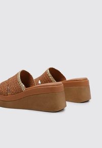 Mules de plataforma trenzados en marrón con lados abiertos, que presentan un upper texturizado y una suela lisa en tono bronceado. Un detalle dorado a lo largo del borde añade un toque de elegancia.