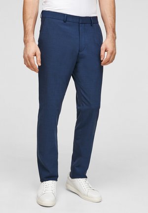 Mann trägt marineblaue Slim-Fit-Hose, weiße Sneakers und ein weißes, eingestecktes Hemd, steht vor einem schlichten hellen Hintergrund.