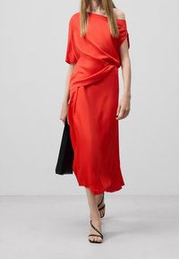 Abito rosso off-the-shoulder in tessuto fluido con dettagli drappeggiati. Accessoriato con una borsa nera e sandali neri flat.