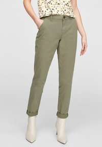 Donna con pantaloni verde oliva arrotolati, stivali alla caviglia bianchi a punta e una canotta floreale, con una mano in tasca.
