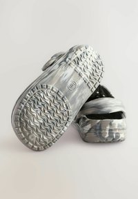 Chaussures à motif camouflage gris avec une semelle en caoutchouc texturée, dos ouvert et sangle ajustable, offrant une surface lisse et une forme arrondie.