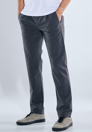 Mann trägt graue Nadelstreifenhose und beige Sneakers mit schwarzen Sohlen, steht mit den Händen in den Taschen vor einfachem Hintergrund.