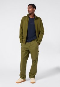 Junger Mann steht mit den Händen in den Taschen, trägt eine olivgrüne Jacke und Hose, ein navyblaues T-Shirt mit Logo und beigen Sneakers.