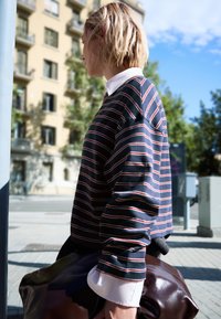 Camicia a maniche lunghe a righe in navy con linee rosse e bianche, con colletto bianco. La persona tiene una borsa marrone lucida, in piedi all'aperto.