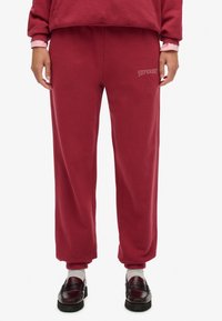 Bordowe spodnie dresowe o luźnym kroju, z elastycznym pasem, mankietami i wyhaftowanym logo "SUPERDRY" po lewej stronie. Miękka, teksturowana tkanina.
