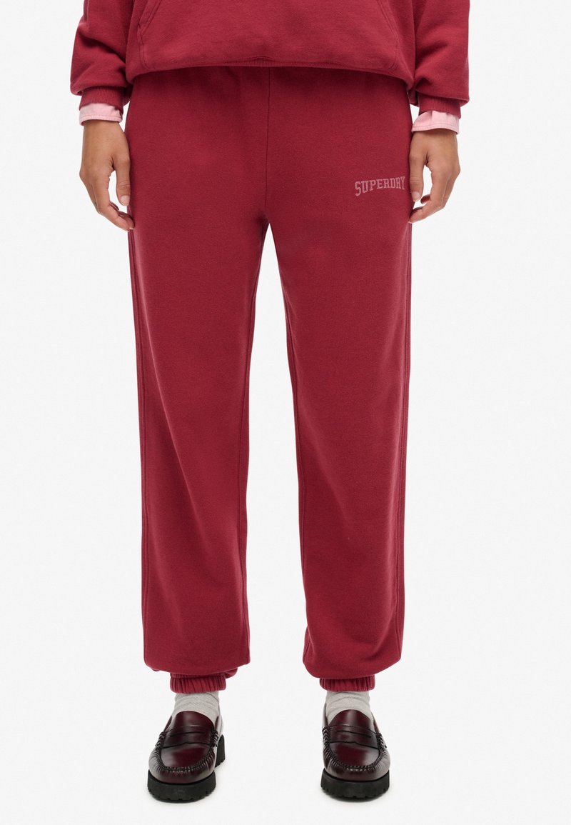 Bordowe spodnie dresowe o luźnym kroju, z elastycznym pasem, mankietami i wyhaftowanym logo "SUPERDRY" po lewej stronie. Miękka, teksturowana tkanina.