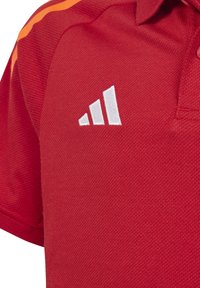 Rode poloshirt met een gestructureerde afwerking, voorzien van het witte adidas-logo op de linkerborst en oranje strepen op de schouders.