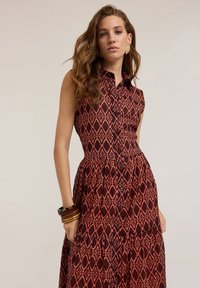 A sleeveless, collared dress featuring a red and black diamond pattern, a fitted bodice, and flowing tiered skirt with button accents.