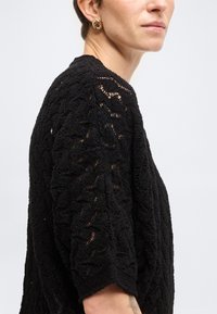 Personne portant un haut en maille noire texturée à manches courtes et une boucle d'oreille circulaire dorée, montrée de l'épaule jusqu'au milieu du visage.
