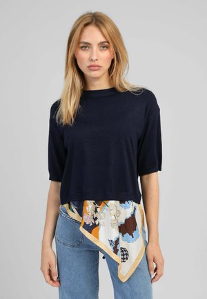 Femme portant un haut bleu marine à manches courtes avec un détail d'écharpe florale multicolore et un jean bleu clair, debout devant un fond gris uni.