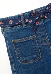 Pantalones vaqueros de mezclilla azul con costuras marrones y un cinturón de tela azul marino con un patrón floral atado en un lazo en la cintura.