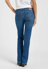 Jeans de mezclilla azul acampanados con una etiqueta azul, bolsillos traseros con costuras decorativas y una cintura alta. El calzado son botas de tacón marrones.