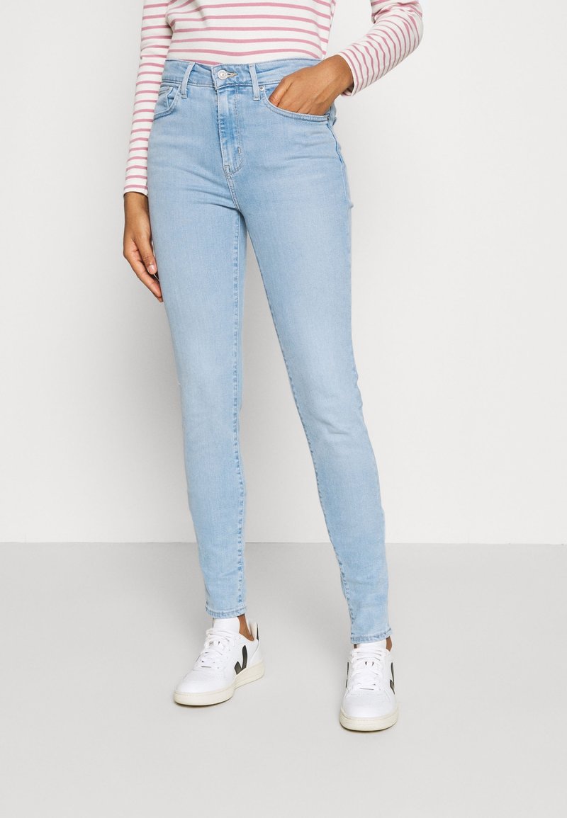 Femme portant un jean skinny bleu clair, des baskets blanches avec un détail noir et un haut à manches longues rayé rose et blanc, debout avec une main dans la poche.