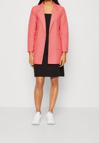 Blazer corallo in tessuto texturizzato, con due tasche frontali e rever a punta; abbinato a un vestito nero aderente e scarpe da ginnastica bianche.