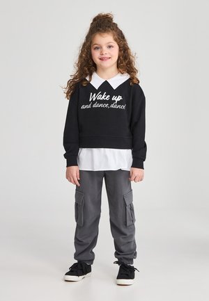 Ragazza giovane con i capelli ricci, che indossa una felpa nera con colletto bianco e scritta, pantaloni cargo grigi e sneakers nere, in piedi e sorridente.