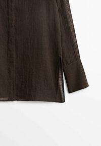 Brązowa koszula z długim rękawem, wykonana z lekkiej tkaniny, o luźnym kroju, z bocznymi rozcięciami i sztywnym kołnierzykiem. Tekstura wydaje się nieco przezroczysta.