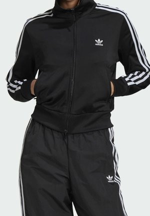 Person iført en sort Adidas træningsdragt med hvide striber på ærmer og bukser, hænderne i jakkelommerne, stående mod en ensfarvet baggrund.