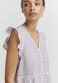 Blouse lavande clair, avec des manches courtes à volants, un décolleté en V profond, une fermeture à bouton et un tissu texturé avec un motif à carreaux subtil.