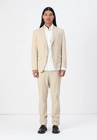 Completo beige con giacca su misura e pantaloni abbinati, in tessuto misto cotone, camicia bianca sotto, abbinato a scarpe nere.