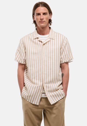 Homme aux cheveux mi-longs portant une chemise beige et blanche à rayures verticales à manches courtes et un pantalon marron clair, mains dans les poches.