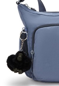 Borsa a spalla in tessuto blu con una texture liscia, dotata di una tasca con zip sulla parte anteriore e un portachiavi a forma di gorilla nero peloso.