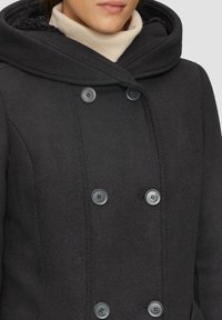 Schwarzer Doppelreiher-Mantel mit strukturiertem Stoff, großem Kapuzen und grauen Knöpfen. Eine kontrastierende Innenauskleidung ist am Kragen sichtbar.