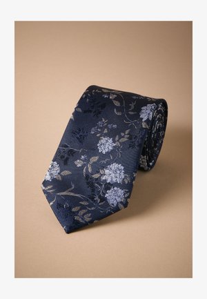 Marineblåt slips med blomsterprint i lyseblå og sølvtoner. Fremstillet af glat stof; har spids ende og rullet form.