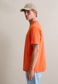 Pomarańczowa bawełniana koszulka z okrągłym dekoltem, krótkimi rękawami i luźnym krojem, zestawiona z jasnoniebieskimi dżinsami i beżową czapką.