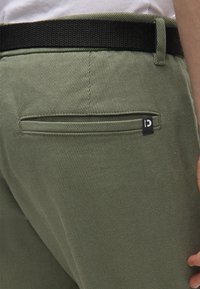 Πράσινο υφασμάτινο παντελόνι με τσέπη στο πίσω μέρος που φέρει μια διακριτική ετικέτα με λογότυπο και μαύρη ελαστική μέση. Κοντινή λήψη από πίσω όψη.